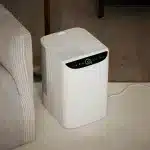 hvit luftfukter på gulv på teppe ved siden av sofa. sort betjeningspanel med grønn ring og hvite knapper